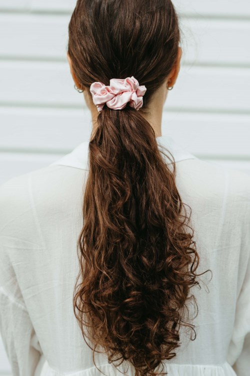 A woman with long, curly brown hair tied with a pink scrunchie, viewed from behind, wearing a light-colored top.