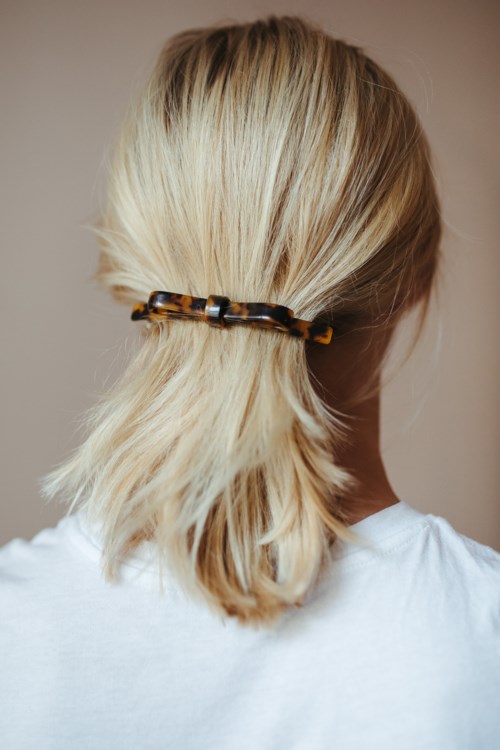 Eine Person hat blonde Haare, die mit einer dunklen Haarklammer zusammengehalten werden, und trägt ein weißes Hemd.
