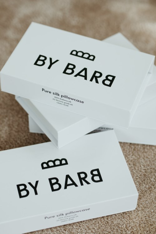 To hvite bokser merket 'BY BARD' inneholder silkesmykkeposer, plassert på en beige overflate.