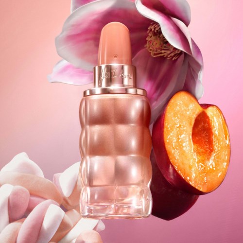 En parfymeflaske omgitt av en blomst, en halvt persikk, og rosa og hvite godterier på en rosa bakgrunn.