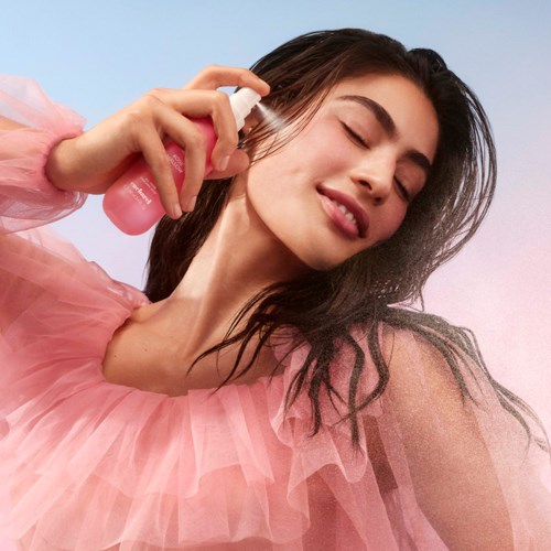 A woman happily sprays perfume on her neck, wearing a pink tulle dress with her eyes closed.