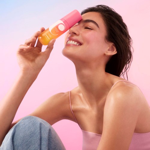 A smiling woman holds a pink and yellow bottle near her face against a pink background.