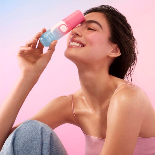 A smiling woman holds a pink and blue spray can near her eye, with a pink background.