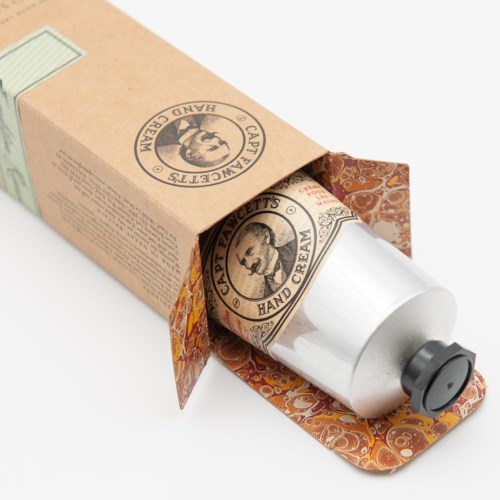 A hand cream tube with a vintage-style label inside a paper box, partially opened. The box has a decorative pattern.