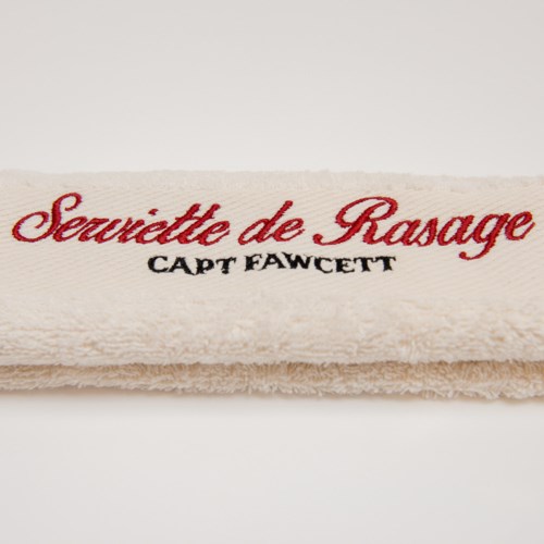 En beige handduk med röd och svart broderad text, läsande 'Se coquettte de Rasage' och 'Capt Fawcett.'