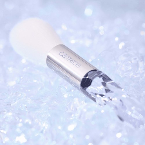 A makeup brush with a white top and a clear handle labeled "catrice" is on icy surface with sparkles.