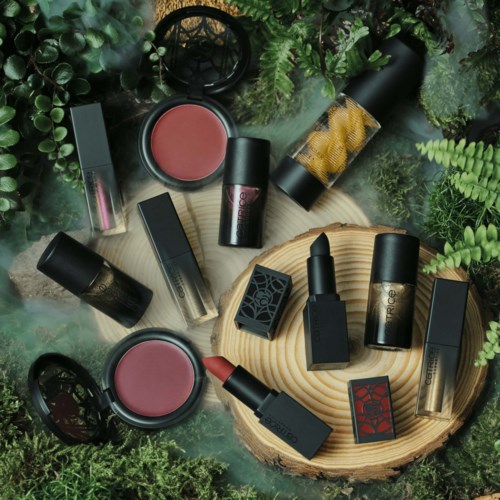 Kosmetikkprodukter som leppestifter, kremer og blush er ordnet på et trestykke omgitt av grønne planter.