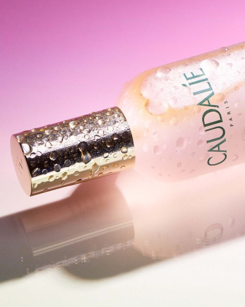 En flaska med spraymunstycke, rosa vätska och vattendroppar, märkt 'CAUDALIE PARIS' på en rosa bakgrund.