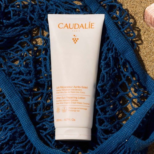 Det er en hvit tube med after-sun lotion omgitt av et blått nett, plassert på sand.