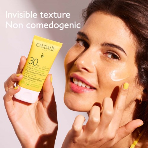 En kvinna som ler och håller i en gul solskyddsdispensare märkt 'Caudalie' med SPF 30.