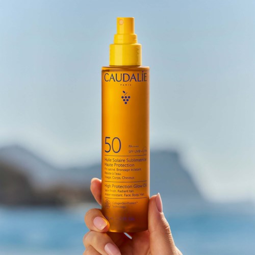En hånd holder en gul flaske med Caudalie solbeskyttelsesspray med SPF 50, utendørs nær vann.