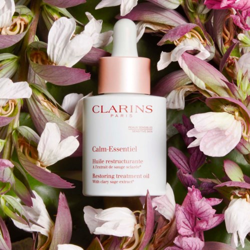 A white bottle of Clarins Calm-Essentiel Restarting treatment oil surrounded by pink, white, and purple flowers.