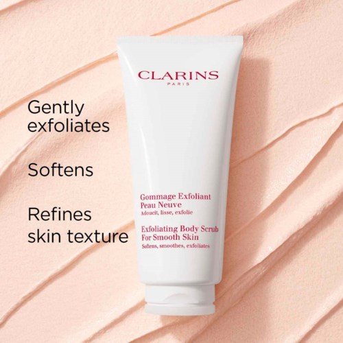En vit tub med Clarins peeling kroppsskrubb med rosa text, placerad på en texturerad persikofärgad bakgrund.