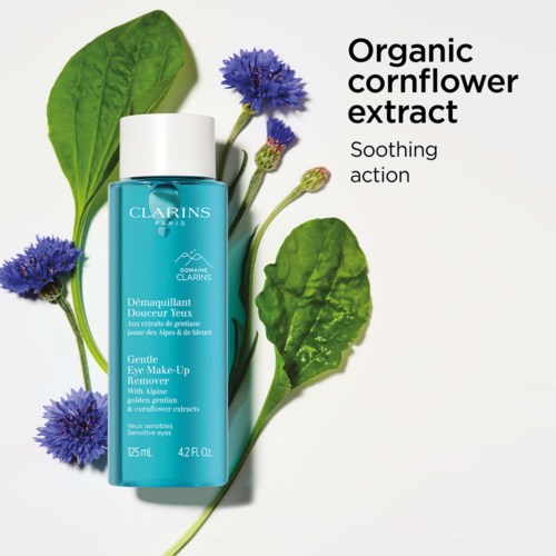 En blå flaska med mild ögonmakeup-remover omringad av gröna blad och blå blommor, med text om ekologiskt kärnkraftsextrakt.