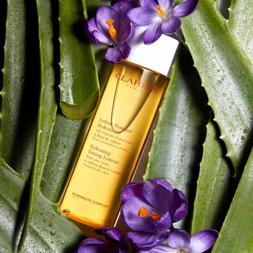 A bottle of Clarins lotion among aloe vera leaves and purple flowers with orange stamens.