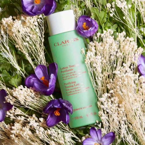 En grön flaska med Clarins-toner omgiven av lila blommor och vita torkade växter.