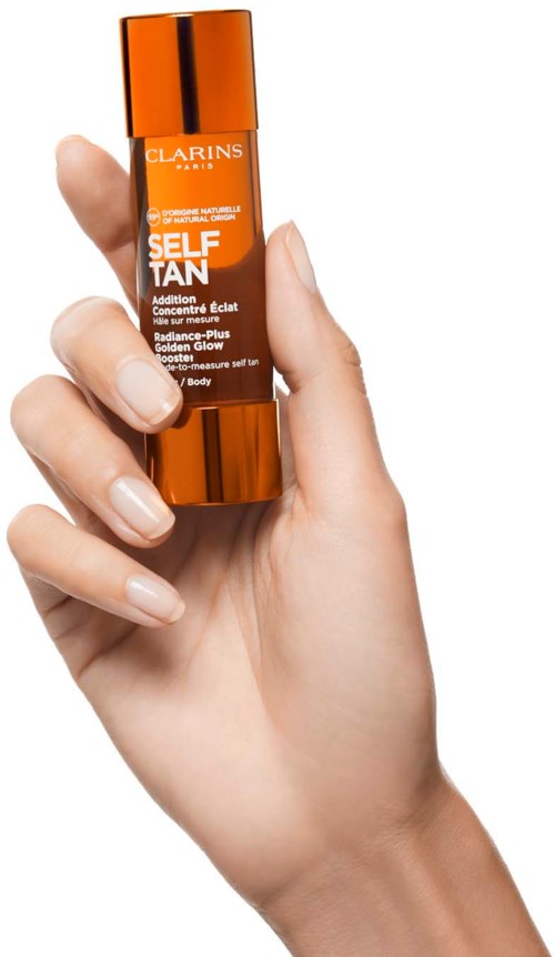 Käsi pitää pientä oranssia pulloa, jonka etiketti on Clarins Self Tanning, ja kansi on kultainen.