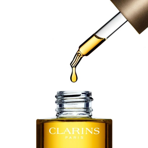 En pipett släpper guldfärgad olja i en liten flaska märkt 'Clarins Paris' på vit bakgrund.