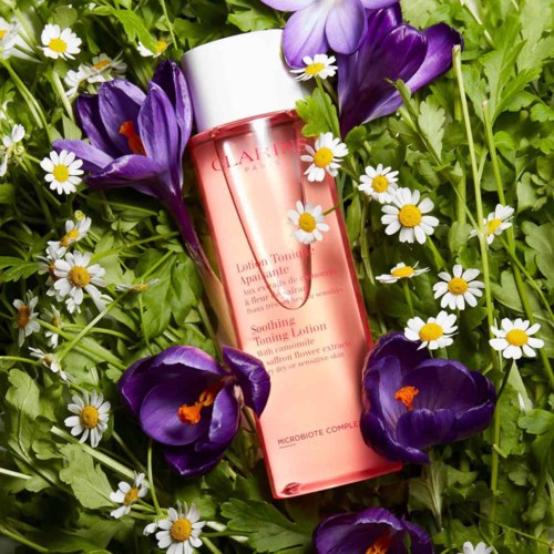 En rosa flaska med Clarins lugnande toningvatten omgiven av lila krokusar och små vita prästkragar i en grön trädgård.