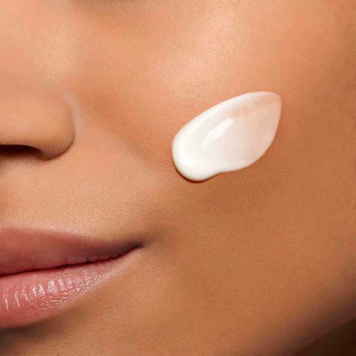 Close-up of a person's face showing a dollop of white cream on cheek. Skin is smooth and makeup-free.