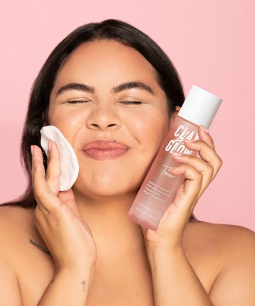 A woman with dark hair holds a pink skincare bottle and uses a cotton pad on her face against a pink background.