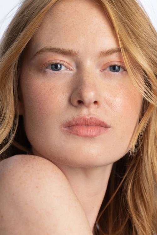 Eine junge Frau mit roten Haaren, heller Hautfarbe, blauen Augen und natürlichem Make-up schaut in die Kamera.