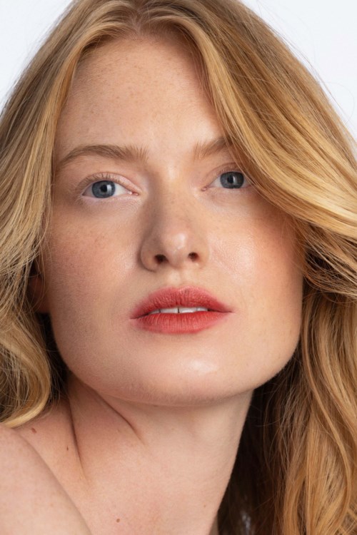 En kvinna med rött hår och blå ögon har ett neutralt uttryck, med mjuk makeup och naturlig hud.