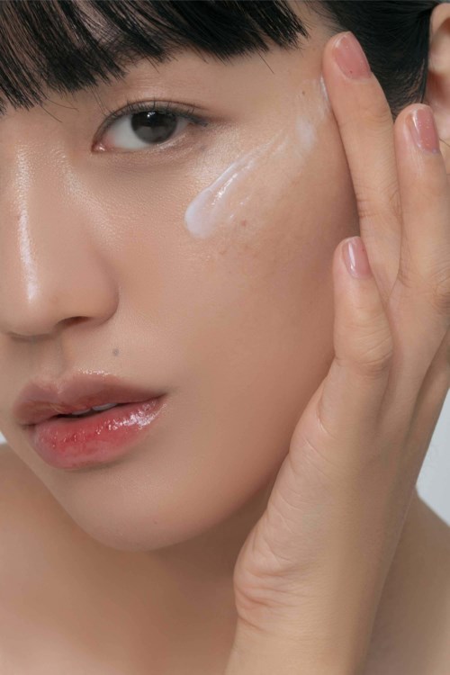 Eine Frau trägt Creme auf oder berührt ihr Gesicht, mit sichtbarer Lotion auf der Wange, in einer Nahaufnahme.