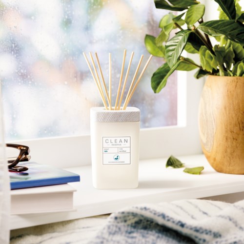 En hvid diffuser med bambusstokke, ved en grøn plante og et vindue, skaber en hyggelig og frisk stemning.