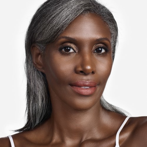 En kvinde med gråt hår og naturlig makeup, iført en hvid top, smiler let. Simpelt baggrund.