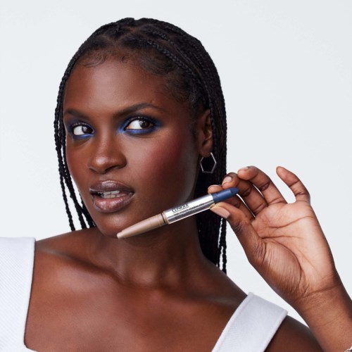 En kvinna med flätat hår och ljus makeup håller en penna nära sitt ansikte, ser självsäker och stilfull ut.