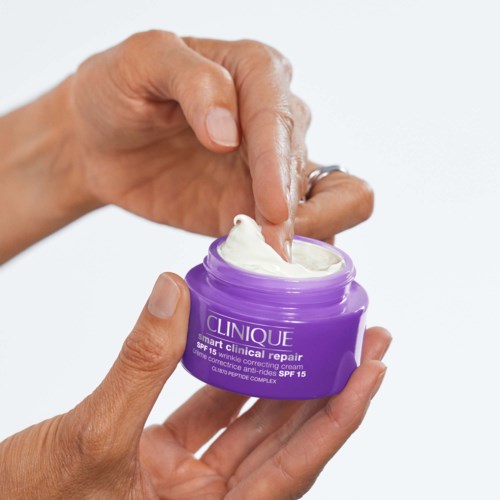 A hand is squeezing white cream from a purple jar labeled Clinique. It is a skincare product for face repair.