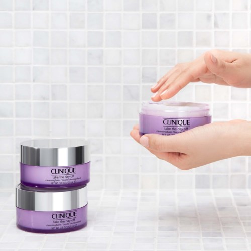 A person is holding a purple jar of Clinique skincare cream, with two more jars stacked below on a tiled surface.