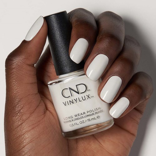 A hand with dark skin holds a white nail polish bottle with black cap, showing painted white fingernails.