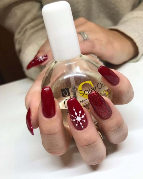Een hand met versierde nagels houdt een fles nagellak met een witte dop. De nagels zijn rood en glinsterend.