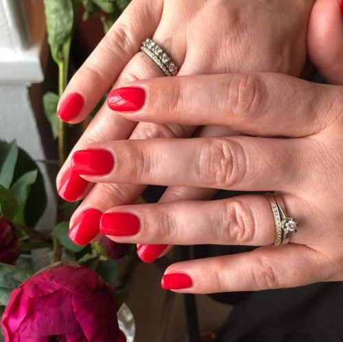 Twee handen met rode nagels, met ringen, voor een bloem en groene planten.