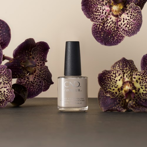 En flaska av tydlig nagellack mellan lila och gula orkidéer på en minimal bakgrund.