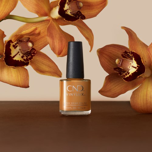En flaska orange nagellack omgiven av orange orkidéer på en mörk yta.