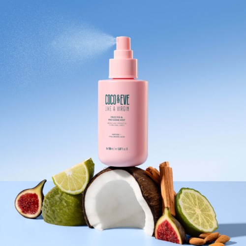 Eine pinke Sprühflasche mit 'COCO&EVE' steht auf Kokosnuss, Limette, Feige, Zimt und Mandelstücken vor blauem Himmel.