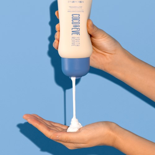 En hånd holder en lotionflaske og trykker creme ud på den anden hånds håndflade mod en blå baggrund.