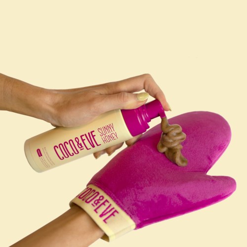 A hand pours a squiggly brown lotion onto a pink glove on a plain background.