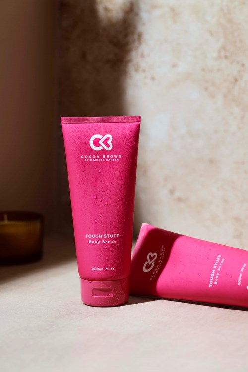 Två rosa flaskor med Cocoa Brown hudvårdsprodukter placerade på en yta, med en upprätt och den andra liggande.