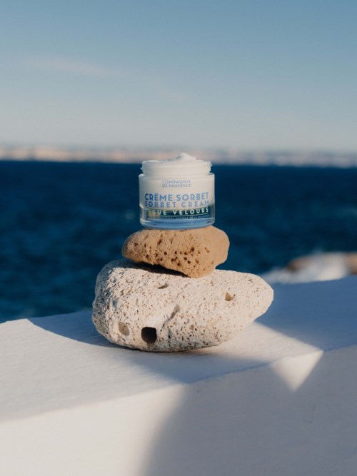 En liten burk med kräm vilar på två stenar vid havet med en klar blå himmel och lugnt vatten i bakgrunden.