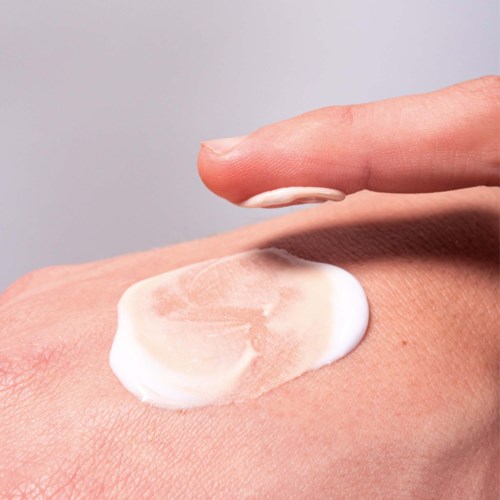 Eine Person trägt weiße Creme mit einem Finger auf die Haut auf vor hellem Hintergrund.