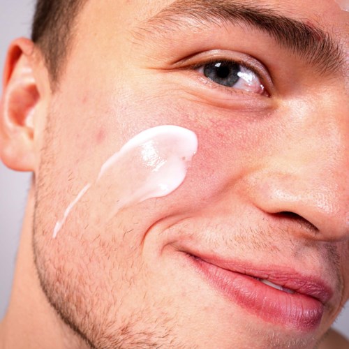 Ein Mann trägt weiße Creme oder Lotion auf die Wange auf, lächelt sanft, Nahaufnahme seines Gesichts.