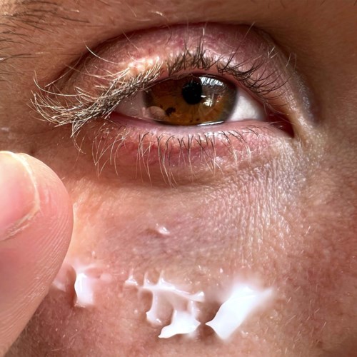 En närbild av ett öga med hud, ögonfransar och ett finger nära den nedre ögonlocket. Någon kräm eller lotion appliceras.