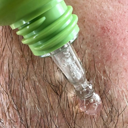 Een groene pipet spuit vloeistof op huid met haar, vormt bellen en een kleine druppel.