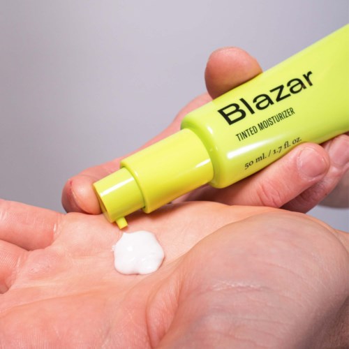 A hand holds a yellow bottle of Blazar tinted moisturizer, with some white cream on the palm.