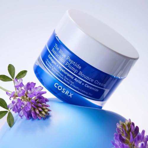 A blue jar of COSRX Blue Peptide Bounce Cream resting on a smooth blue surface with purple flowers nearby.