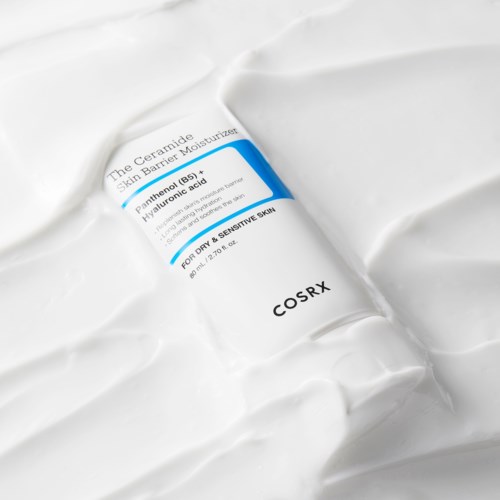 A white tube labeled COSRX with blue and black text, lying on a textured white surface.